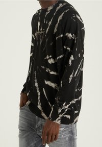 Sudadera negra con estampado de tie-dye blanco, cuello redondo, mangas largas y puños de canalé. Combinada con jeans gris claro.