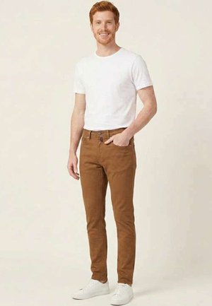 Jeune homme aux cheveux roux portant un t-shirt blanc, un pantalon marron et des baskets blanches, debout avec une main dans la poche.