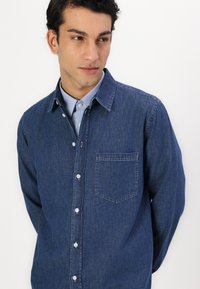 Denim overhemd in donkerblauw met een button-down kraag, een borstzak en witte knopen. De stof is glad met een licht vervaagde textuur.