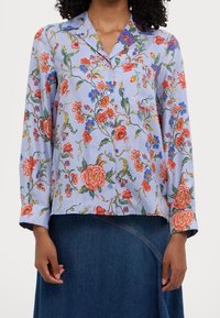 Blouse florale en tissu bleu clair avec des manches longues, ornée de motifs floraux rouges et bleus, d'un col et d'une poche poitrine.