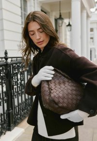 Barna szövött bőrtáska fekete kabát alatt tartva, fehér kötött kesztyűkkel. A texturált táska részletes fonással gazdagított, ami fokozza a dizájnját.