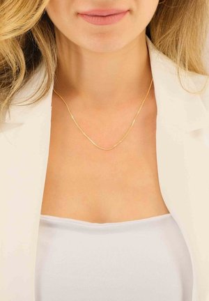 Collier en or avec un design lisse et délicat, présenté sur un décolleté nu, accompagné d'un haut blanc et d'un blazer.