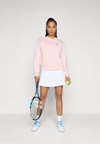 Kvinne i rosa sweatshirt og hvit tennis-skjørt som holder tennisracket og ball, står foran en ensfarget bakgrunn.