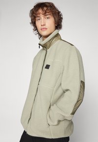 Giacca in fleece di colore beige chiaro con colletto alto, chiusura con zip, tasche laterali e dettagli in verde oliva sulle spalle. Finitura in tessuto testurizzato.