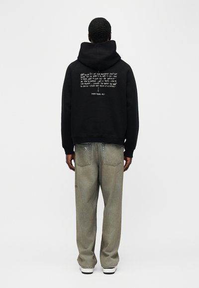 Άνδρας που φοράει μαύρο φούτερ με κουκούλα, με χειρόγραφο λευκό κείμενο στην πλάτη και φαρδιά γκρι τζιν, στέκεται μπροστά σε φωτεινό απλό φόντο.