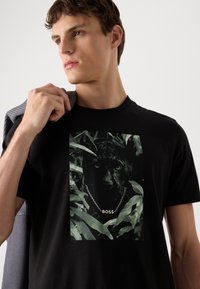 Zwarte t-shirt met een afbeelding van een panter omringd door jungleplanten. De panter draagt een zilveren ketting met de tekst "BOSS". Korte mouwen, katoenen materiaal.