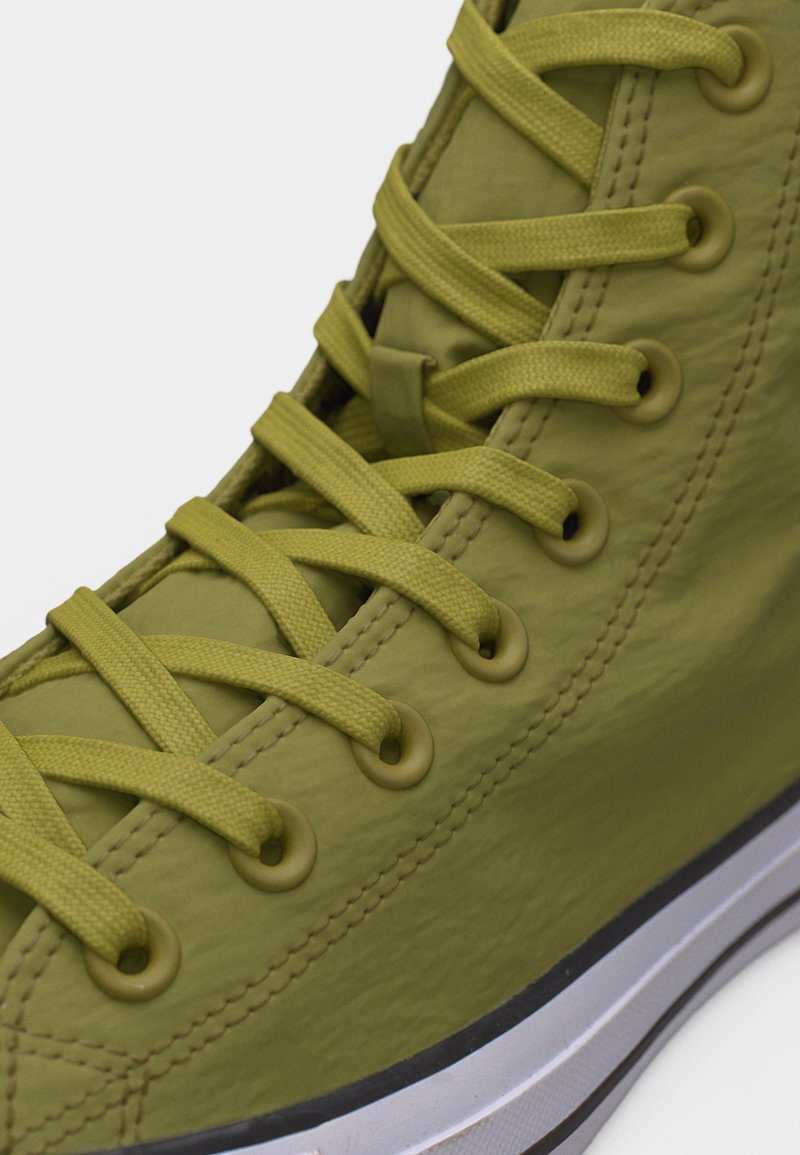 Primer plano de una zapatilla alta de color verde oliva mostrando los cordones, ojales metálicos, costuras cosidas y suela de goma blanca con una franja negra.