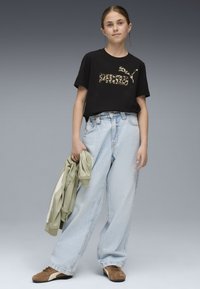 Črna bombažna majica s leopardjim potiskom, svetlo modri oversize jeans in rjave superge. Drži svetlo zeleno jakno.