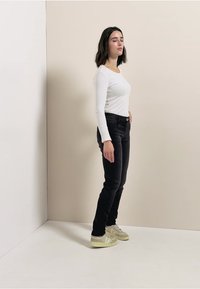 Vit långärmad skjorta, svarta jeans och ljusa sneakers. Stående pose mot en neutral bakgrund med en enkel, ren estetik.