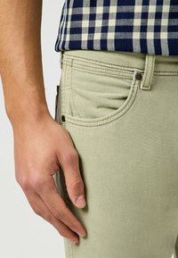 Jeans verde claro con una textura suave, que presentan un bolsillo delantero y costuras marrones. La mano está situada cerca de la zona del bolsillo.