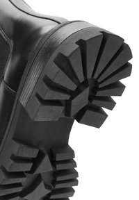 Suela de bota de goma negra con un patrón de banda de rodadura resistente, con profundos surcos y formas angulares para mejorar la tracción y durabilidad.