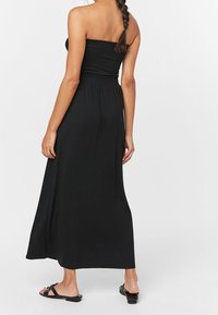 Robe maxi noire sans bretelles avec un corsage ajusté et une taille élastique, présentant une texture lisse et une silhouette fluide. Portée avec des sandales noires.