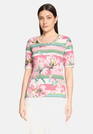 Kobieta w krótkim rękawie koszuli w kwiaty z różowymi, zielonymi i białymi paskami oraz dużymi różowymi kwiatami, w połączeniu z białymi spodniami.