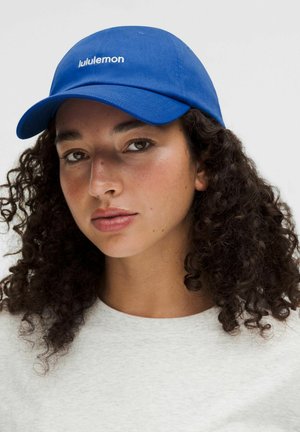 Joven con cabello oscuro y rizado, lleva una gorra azul de Lululemon y una camisa gris clara, mirando directamente al frente con expresión neutral.