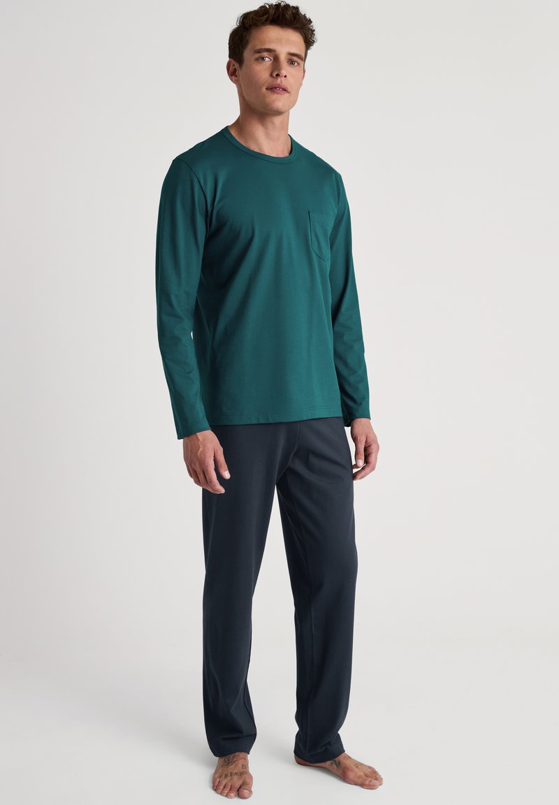Lange mouwen, teal shirt met een borstzak, gecombineerd met donkerblauwe broek. De stof lijkt zacht met een gladde textuur, geschikt voor loungewear.