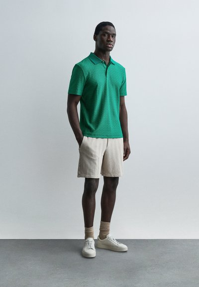 Άνδρας στέκεται με τα χέρια στις τσέπες, φορώντας πράσινο πικέ πόλο πουκάμισο, μπεζ σορτς με σχέδια, μπεζ κάλτσες και λευκά αθλητικά παπούτσια μπροστά σε απλό φόντο.