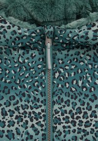 Futboliukas su teal leopardų raštu. Sukurtas su užtrauktuku, minkšta dirbtine vilna viduje ir struktūrizuotu gobtuvu. Tekstūruotas audinys kontrastuoja su glotniu užtrauktuku.