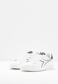 Bílé tenisky s hladkým koženým povrchem, černými akcenty, perforovanými detaily a gumovou podrážkou. Obsahují šněrovací design a logo na jazyku.