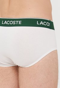 Herren weiße Baumwollslips mit einem grünen elastischen Bund, der ein weißes "LACOSTE"-Logo zeigt. Glatte Textur und enganliegendes Design.