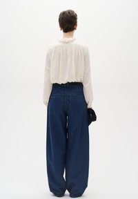 Krémová blůza s volánovým límcem, spárovaná s oversized tmavě modrými džíny. Zadní pohled ukazuje volný střih a dvě zadní kapsy.