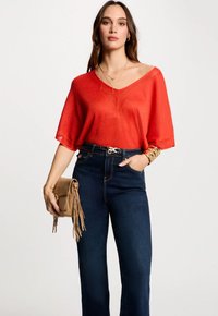 Rode losse gebreide top met V-hals, gecombineerd met hooggesneden donkerblauwe jeans. Accessoires zijn een kleine tan geknoopte tas en gelaagde kettingen.