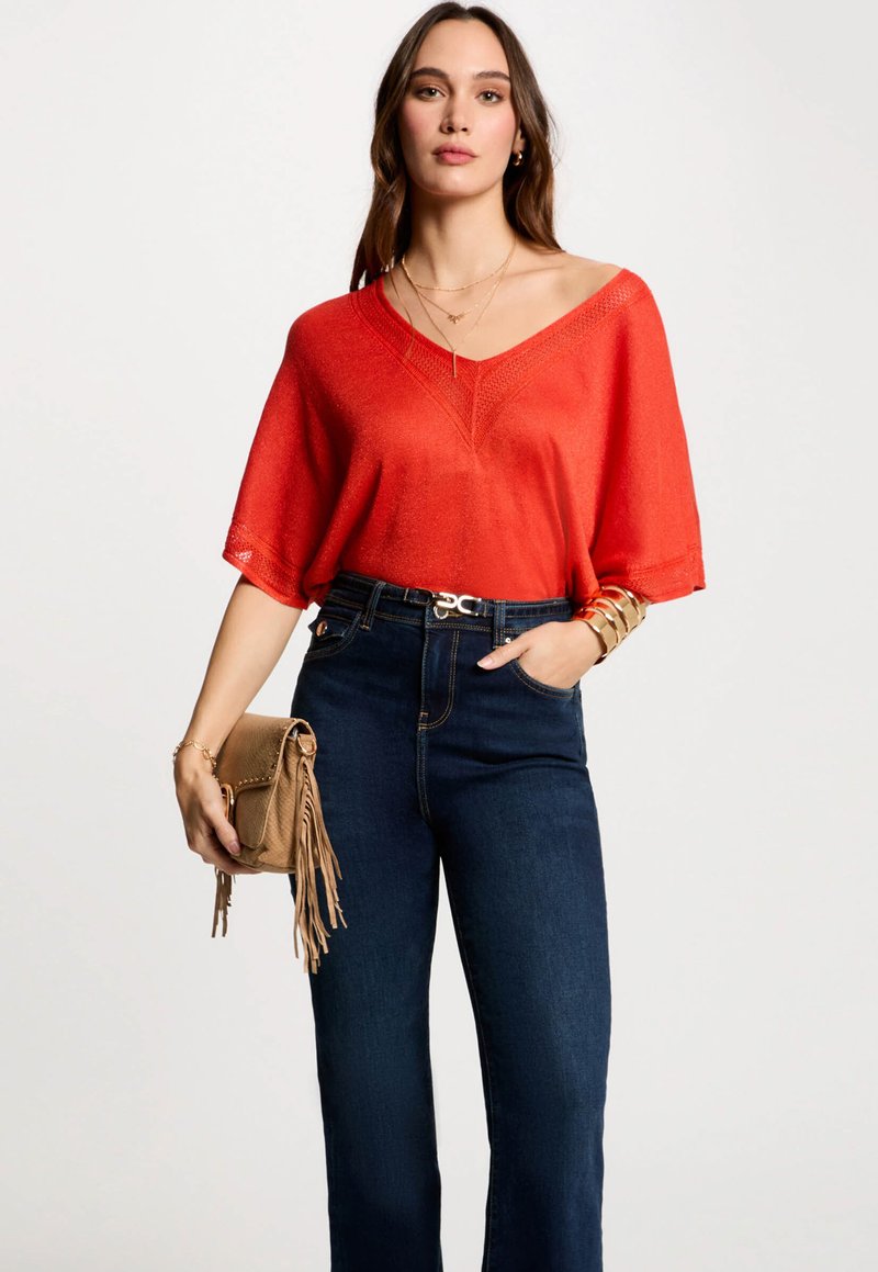 Rode losse gebreide top met V-hals, gecombineerd met hooggesneden donkerblauwe jeans. Accessoires zijn een kleine tan geknoopte tas en gelaagde kettingen.