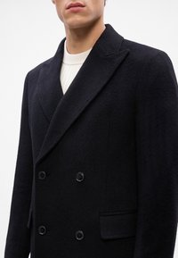 Cappotto nero in lana doppiopetto con ampi risvolti, tessuto testurizzato e due tasche frontali. Presenta bottoni neri sul davanti.