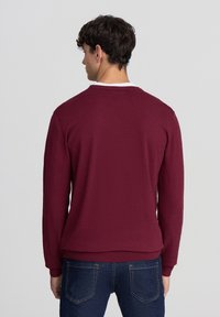 Maglione a maglia bordeaux con maniche lunghe, scollo a giro e polsini a coste. Indossato sopra una camicia bianca, abbinato a jeans in denim blu.