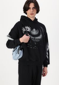 Schwarzer oversized Hoodie mit grafischem Design und Logo, kombiniert mit einer kleinen hellblauen Umhängetasche mit Reißverschlüssen und Klappenakzenten.