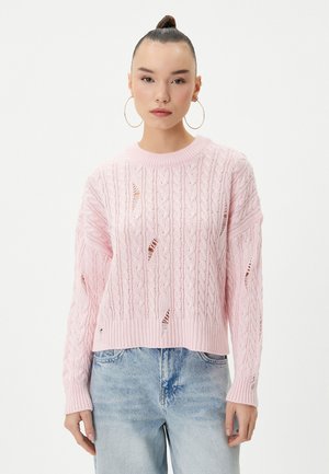 Rosa stickad tröja med ett kabelmönster, med stora utskärningar och ribbade ärmslut. Kombinerad med ljusblå denimjeans.
