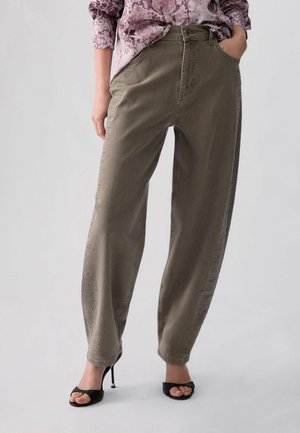 Donna con pantaloni a gamba larga verde oliva con dettagli laterali sottili, una camicetta viola fantasia e sandali neri con tacco alto.