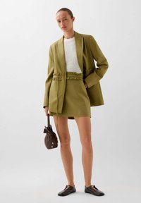 Olijfgroen blazer met gerimpelde taille en zakken, gecombineerd met een gebreid wit topje en een bijpassende olijfgroene rok. Bruine trekkoordtas en zwarte schoenen.