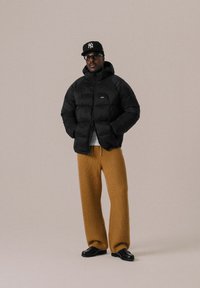 Doudoune noire avec fermeture éclair à l'avant et logo, associée à un pantalon large et texturé marron. Les accessoires comprennent des lunettes de soleil noires et une casquette.