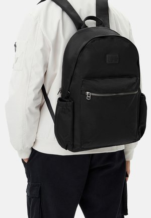 Osoba nosící černý batoh s přední kapsou na zip a bočními přihrádkami, oblečená v bílé bundě a černých kalhotách, stojící s rukama v kapsách.