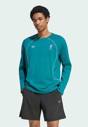Junger Mann trägt ein langärmliges Liverpool FC Shirt in Teal und schwarze Adidas-Shorts, steht mit den Händen in den Taschen vor einem schlichten Hintergrund.