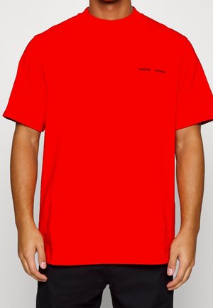 Mand iført en knald rød kortærmet T-shirt med lille sort "SAMSØE" logo på venstre bryst, kombineret med sorte bukser, mod en ensfarvet baggrund.