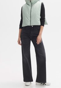 Personne portant un gilet matelassé court vert clair, une chemise noire à manches longues, un jean foncé à jambes larges et des baskets blanches avec des rayures noires.
