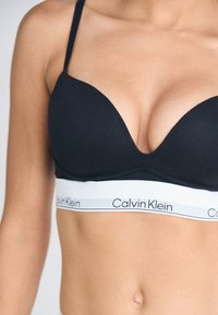 Black padded bra with plunge design, featuring a white elastic band with "Calvin Klein" logo. Adjustable straps and smooth texture.