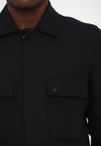 Schwarze, gewebte Jacke mit Kragen, vorderem Knopfverschluss, zwei Brusttaschen und einem kleinen metallischen Logo-Detail auf der linken Tasche.