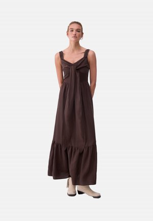 Donna in piedi con le mani dietro la schiena che indossa un vestito lungo marrone scuro senza maniche con dettaglio di fiocco davanti e stivaletti beige.