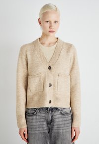 Μπεζ πλεκτό πουλόβερ με V λαιμόκοψη, ριχά μανίκια και δύο εμπρόσθιες τσέπες; διαθέτει μεγάλα σκούρα κουμπιά και απαλό υφάσματος.