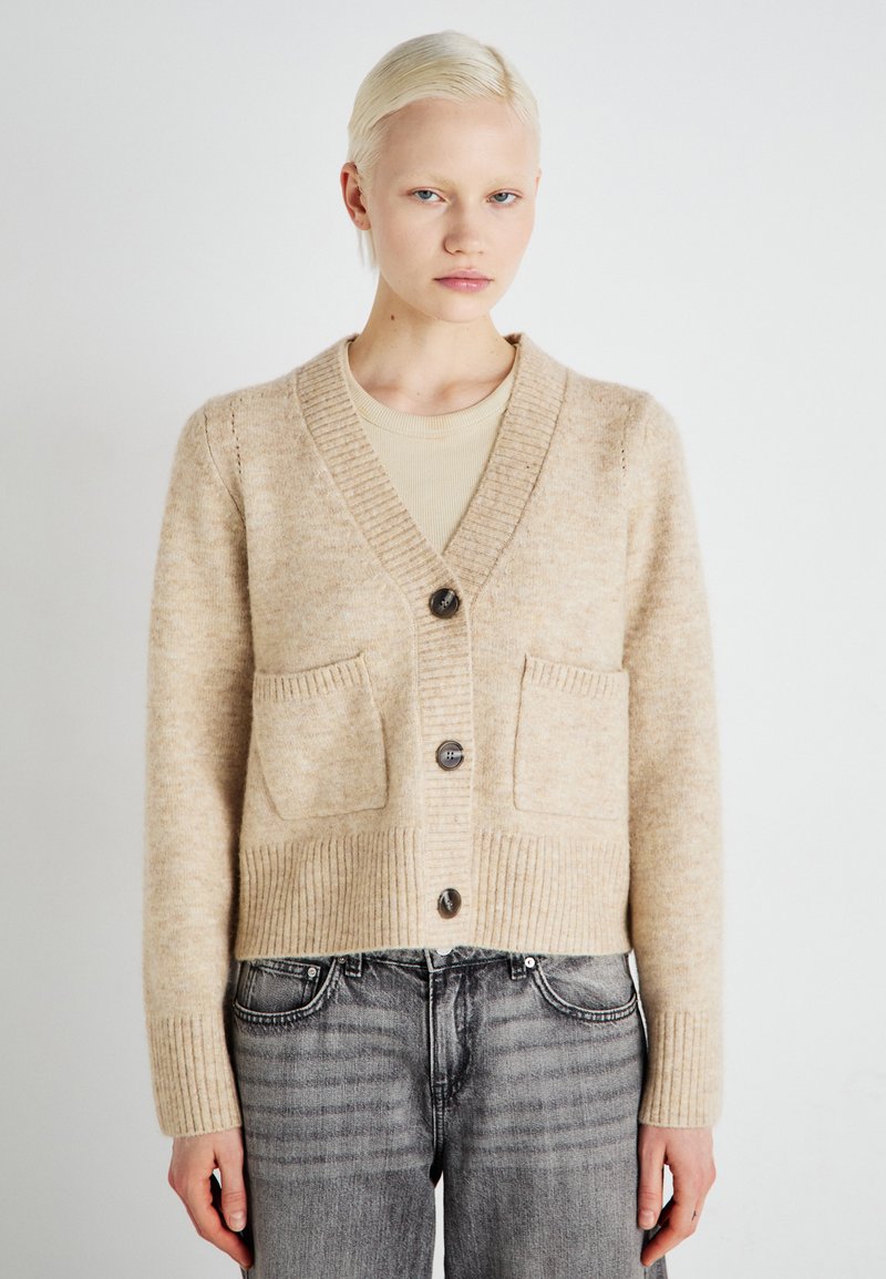 Μπεζ πλεκτό πουλόβερ με V λαιμόκοψη, ριχά μανίκια και δύο εμπρόσθιες τσέπες; διαθέτει μεγάλα σκούρα κουμπιά και απαλό υφάσματος.