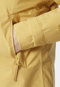 Chaqueta impermeable amarilla con una textura suave, que cuenta con un bolsillo con cremallera y un acento de logo en la manga. Diseño ajustado.