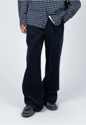 Persoon die losse donkere corduroy broek, zwarte glanzende schoenen en een marineblauw geruit overhemd draagt met handen in de zakken.