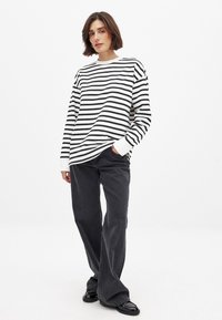 Bela srajca z dolgimi rokavi in črnimi črtami, v kombinaciji z ohlapnimi sivimi hlačami. Srajca ima sproščen kroj s krožnim izrezom in rebrastimi manšeti.