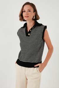 Žena s kratkom smeđom kosom, koja nosi zlatne naušnice, crno-bijeli prugasti bezrukavni džemper s ovratnikom i svijetlo bež hlače, ruke u džepovima.