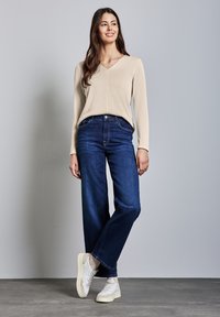 Beige långärmad V-ringad tröja, blå vida jeans och vita sneakers. Modellen står mot en grå bakgrund.