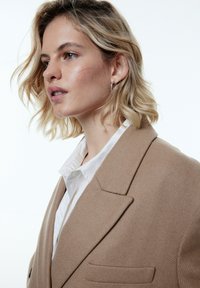 Beige wollen blazer met ingezette revers, voorzakken en een zachte textuur, gedragen over een wit overhemd met kraag. Lichaam gezien in profiel.