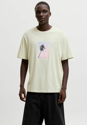 Mann trägt hellbeiges T-Shirt mit Palmenmotiv und schwarze Hose, steht vor einem einfarbigen hellen Hintergrund.