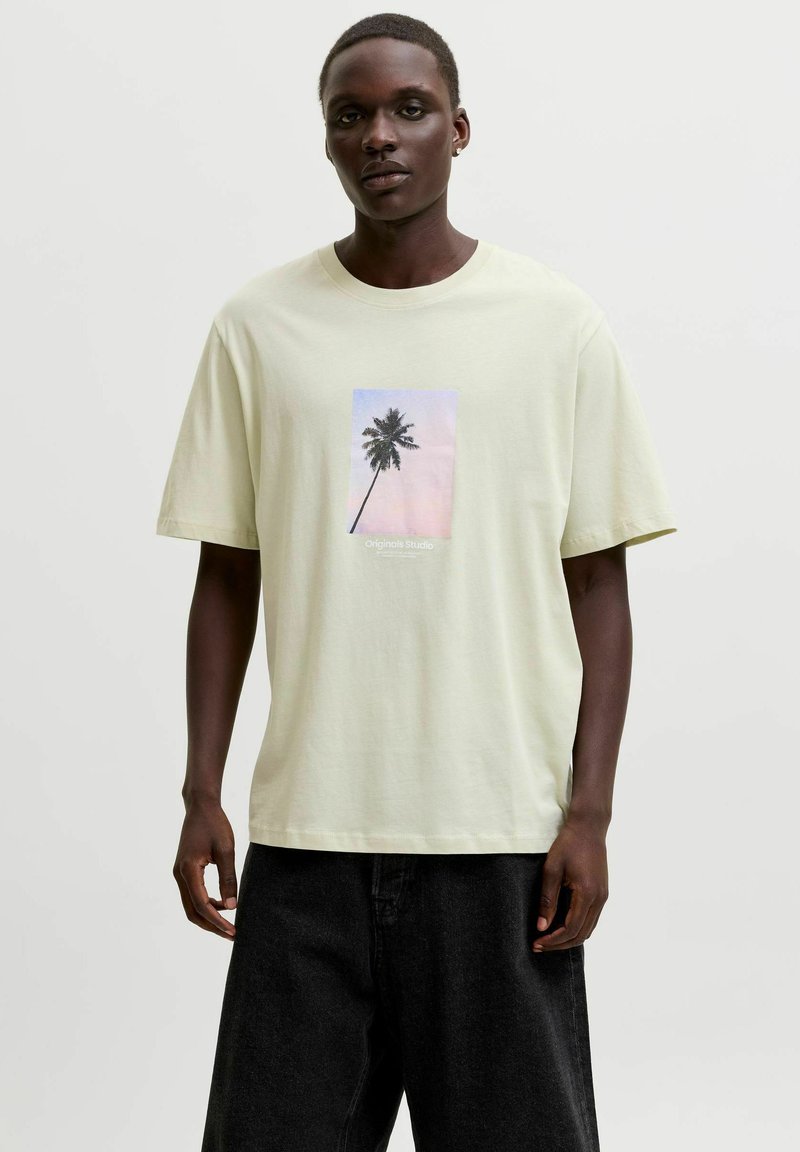 Homme portant un T-shirt beige clair avec un motif de palmier et un pantalon noir, debout devant un fond clair uni.
